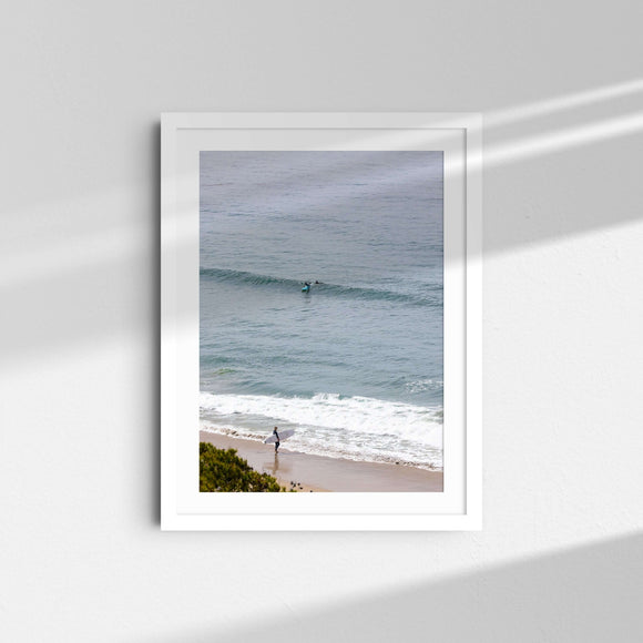 A framed fine art photography print featuring a surfer heading into the calm, light blue ocean in Malibu, California.