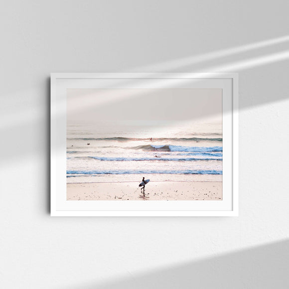 A framed fine art photography print featuring a surfer walking with a surfboard in front of a pink and purple sunset over the ocean in Malibu, California.