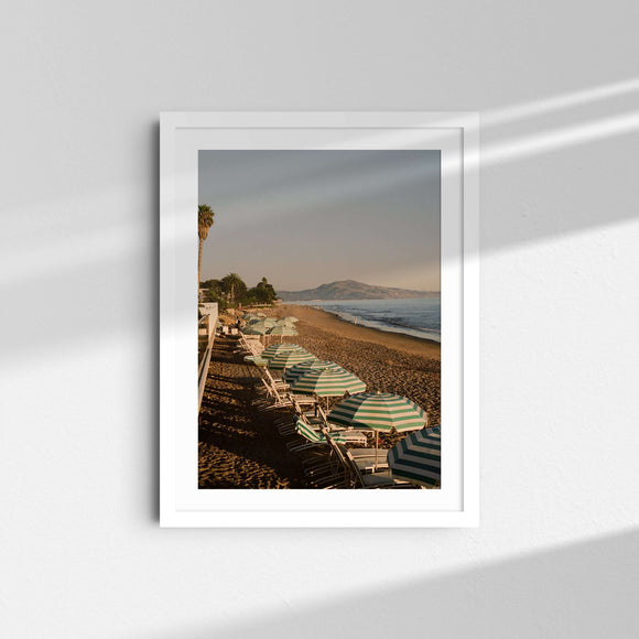 A framed fine art photography print featuring the sunset and blue umbrellas lining the beach in Santa Barbara, California.