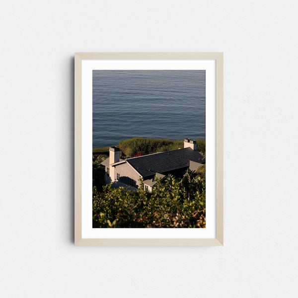 A framed fine art photography print featuring a gray shingled house overlooking blue ocean.