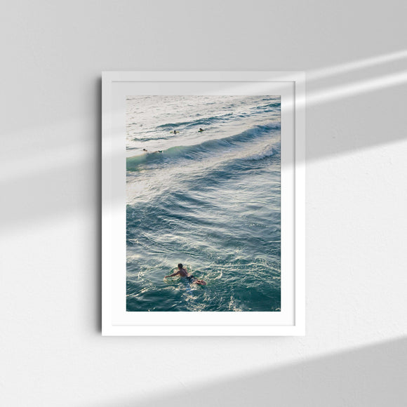 A framed fine art photography print featuring a surfer swimming through the deep blue waters.