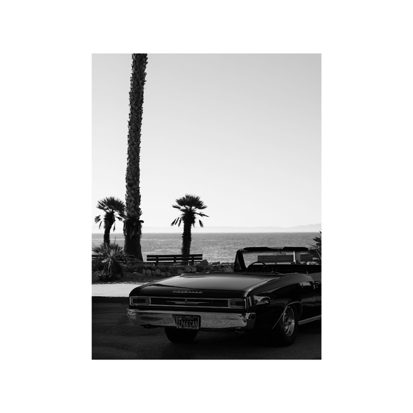 A framed fine art photography print featuring a black and white image of a vintage car in front of the ocean and palm trees.