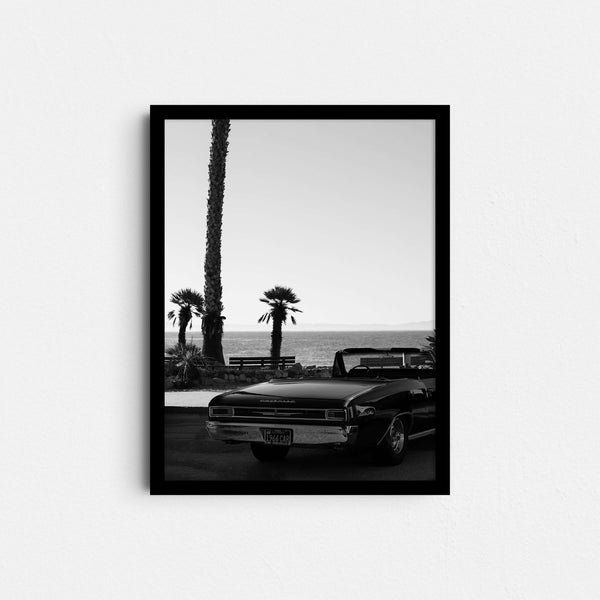 A framed fine art photography print featuring a black and white image of a vintage car in front of the ocean and palm trees.