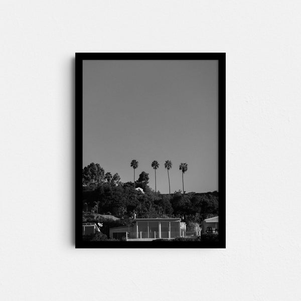 A framed fine art photography print featuring a black and white photo of palm trees and a house in the hills of Malibu, California.