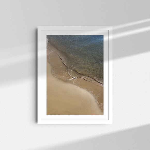 A framed fine art photography print featuring the curve of an ocean wave against the sand.